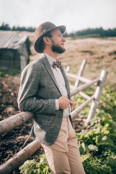 Young man in stylish clothes and hat stands on background of old wooden hut and fence. - Foto, Bild