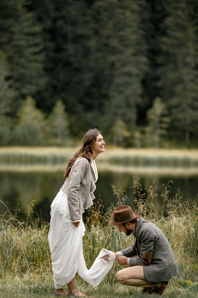 Groom cleans dirty hem of bride's dress and they laugh happily near forest lake. - Photo, image