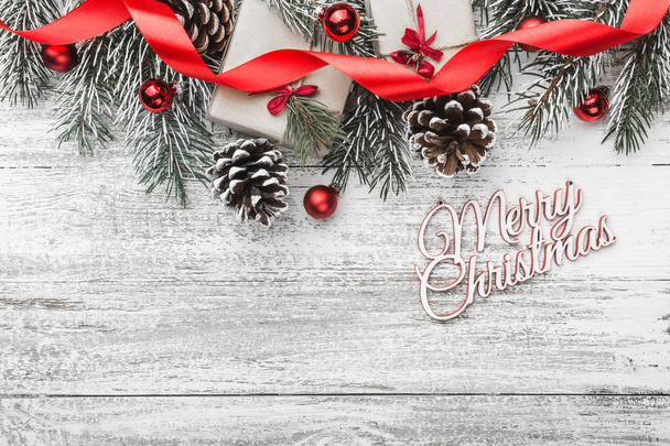 Vista desde arriba, arriba, sobre un fondo de tabla de madera blanca con rama de abeto con pinos, cajas de regalo, cinta, globos de árbol rojo e inscripción Feliz Navidad con espacio para texto, tarjeta de felicitación
 - Foto, imagen