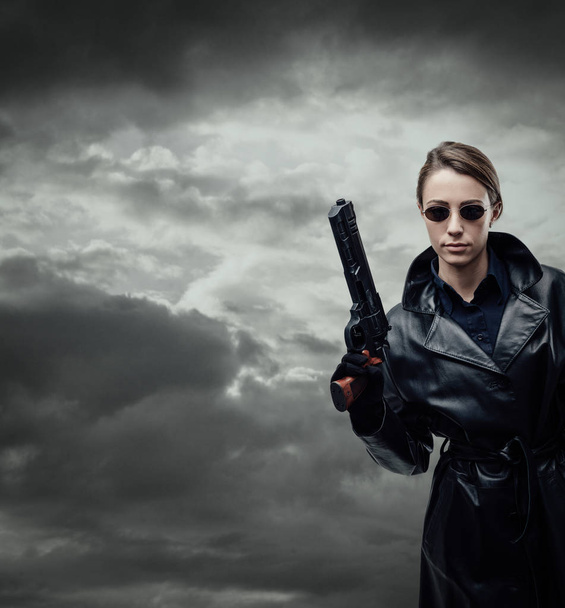 Cool female spy agent in black leather coat, she is holding a gun and posing - Foto, Bild