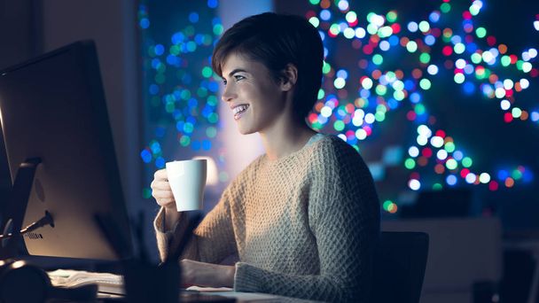Smiling young woman connecting late at night with her computer and having an hot drink, she is watching movies online - Foto, afbeelding