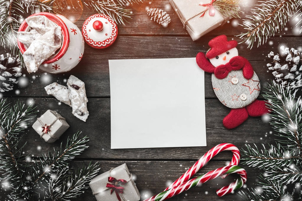 Weihnachtsthema mit Teedose, Tannenzweigen, Schneemannspielzeug, Zapfen, Geschenkschachteln und Bonbons, Notizblock und Stift für Text, auf Holzgrund von oben betrachtet, im Schnee, Grußkarte  - Foto, Bild
