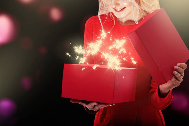 una giovane bionda con un vestito rosso sta strappando una confezione regalo. Dalla scatola c'è una luce brillante e stelle
. - Foto, immagini