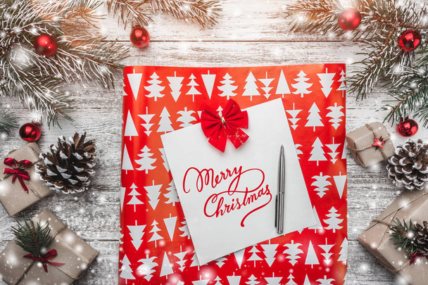 View from above, of a paper sheet and pen with Merry Christmas sign, on white wooden background, with fir branches, red globes, present boxes, cones, greeting card  - Photo, Image