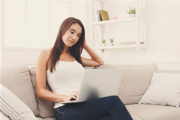Young women using laptop sitting on sofa in white livingroom - Foto, afbeelding