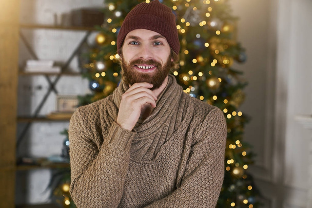 Young hipsters with fashionable beard ponders what to give for Christmas or new year his girlfriend or friend on the background of Christmas tree. Cozy apartments with xmas design. loft party concept. - 写真・画像
