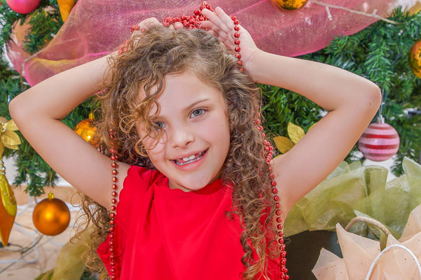 Close up of happy girl wearing a red dress and holding a christmas decoration in her hands over her head, with a Christmas tree behind, christmas concept - Foto, immagini