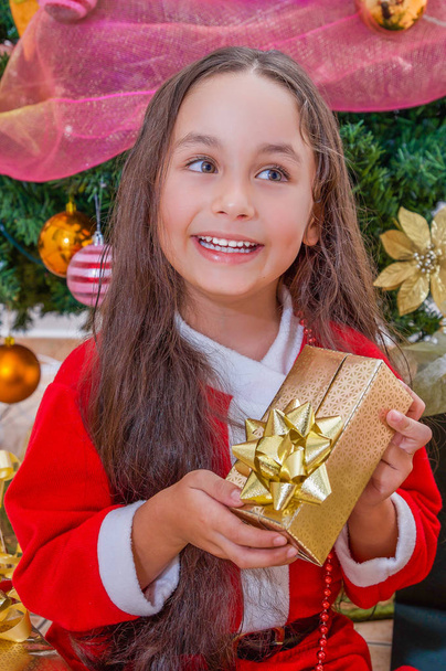 Close up of smiling girl wearing a red santa costume and holding a gift in her hands, with a Christmas tree behind, christmas concept - Фото, зображення