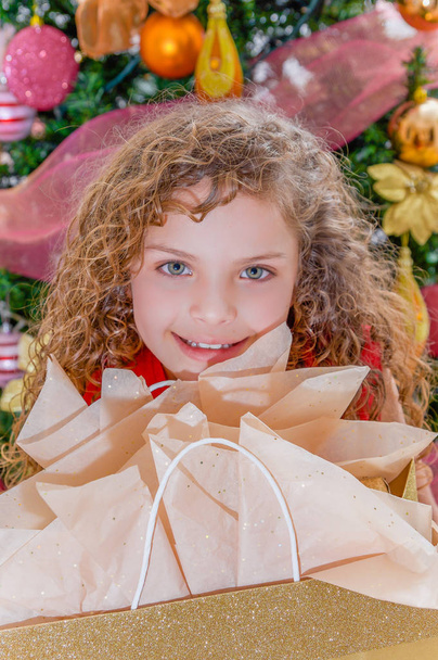 Close up of smiling beautiful curly girl wearing a red blouse and holding a gift in her hands, with a christmas tree behind, christmas and new year concept - Foto, immagini