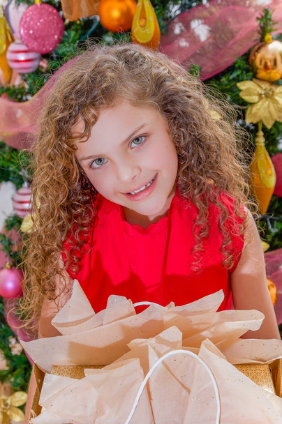 Nahaufnahme eines lächelnden, schönen, lockigen Mädchens, das eine rote Bluse trägt und ein Geschenk in der Hand hält, mit einem Weihnachtsbaum im Hintergrund, Weihnachts- und Neujahrskonzept - Foto, Bild