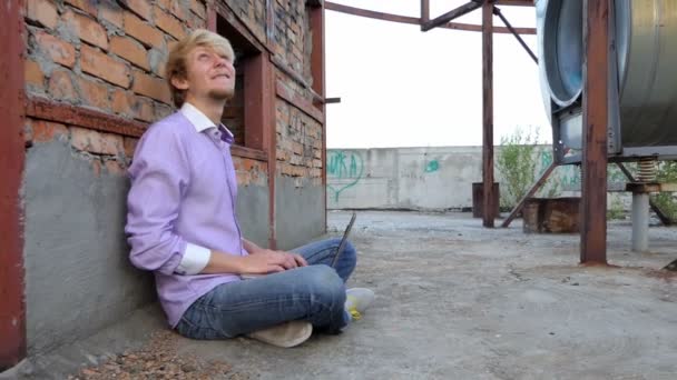 Smiling man sits on a construction site and works at his laptop - Záběry, video