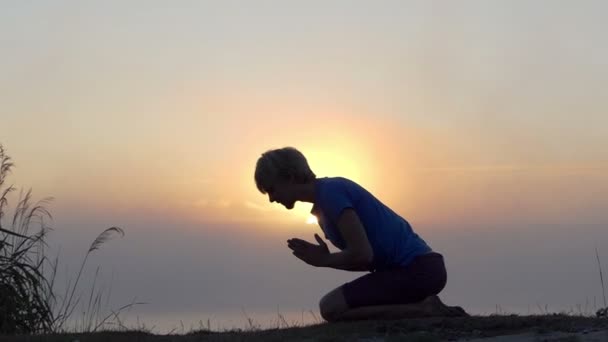 A fiatalember térdel, és imádkozik a tó partján, napnyugtakor a slo-mo - Felvétel, videó
