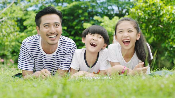 Aasialainen perhe etsii ylös ja nauraa, kun makaa vatsassa ja lukee nurmella
 - Valokuva, kuva