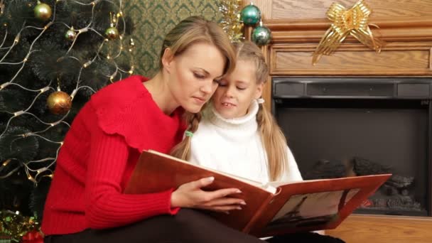 母亲给她的女儿在一棵圣诞树下读一个故事。读一个故事. - Πλάνα, βίντεο