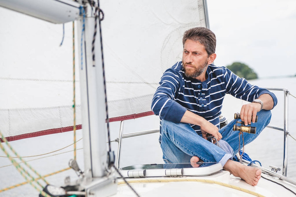 man sits on bow of sailing yacht and holds binoculars - 写真・画像