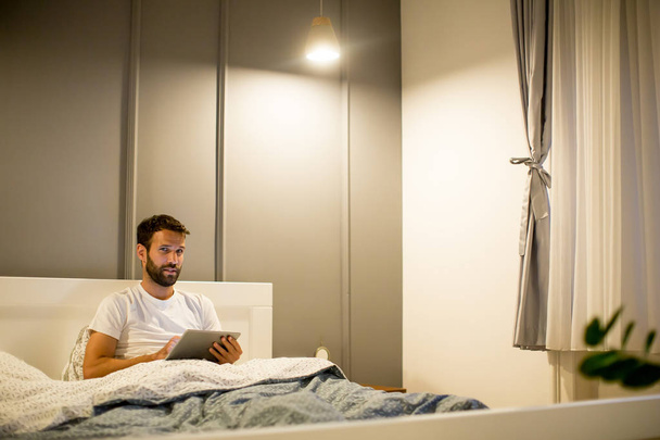 Młody człowiek za pomocą cyfrowego tabletu leżąc na łóżku - Zdjęcie, obraz