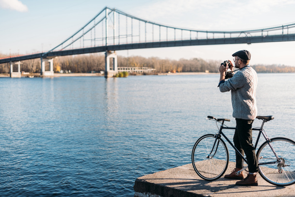 όμορφος άνδρας με vintage φιλμ φωτογραφικής μηχανής και ποδηλάτου στέκεται στην όχθη του ποταμού - Φωτογραφία, εικόνα
