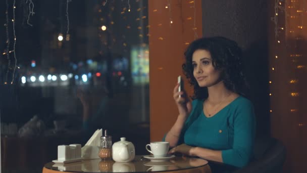 Menina com um smartphone em um café.Uma menina bonita está sentada em um café e falando ao telefone. Ela sorri. Fora da janela ilumina a cidade da noite
. - Filmagem, Vídeo