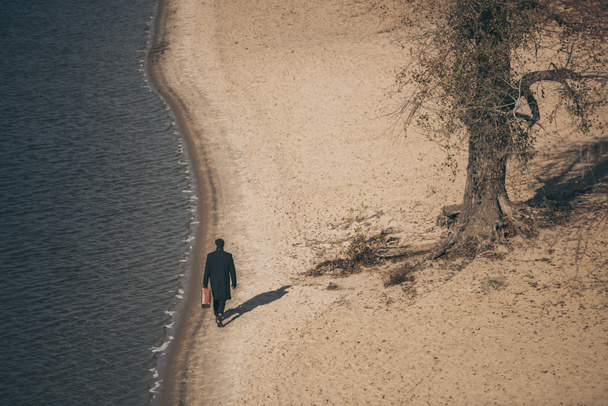vista de ángulo alto del hombre con maleta caminando por la playa de arena en otoño
 - Foto, imagen