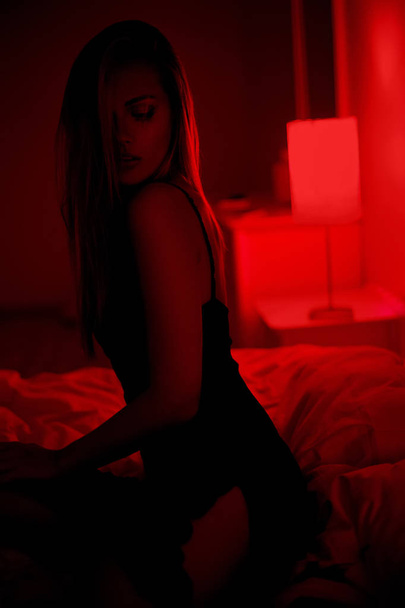 Dark photo of sexy beautiful brunette woman sitting on bed in black dress posing in red interior - Foto, Imagem