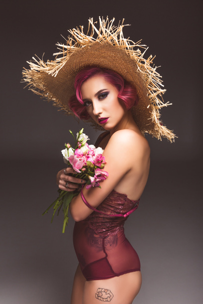 αρκετά ροζ μαλλιά σγουρά κορίτσι σε ψάθινο καπέλο που ποζάρει με λουλούδια μπροστά από γκρι φόντο  - Φωτογραφία, εικόνα