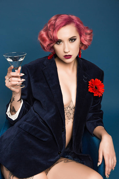 beautiful tattoed pin up girl in jacket with boutonniere and wineglass infront of blue background - Fotó, kép