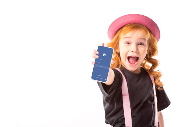 stylish kid in pink hat and suspenders showing smartphone with facebook page isolated on white - Photo, Image