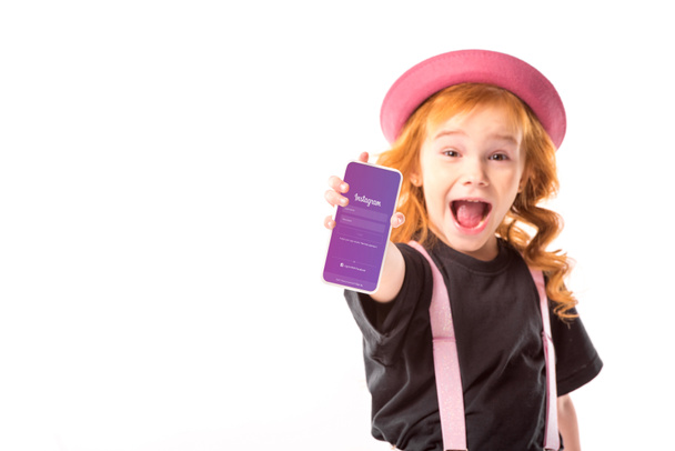 stylish kid in pink hat and suspenders showing smartphone with instagram page isolated on white - Фото, зображення