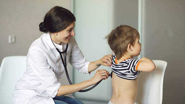 Nuori nainen lääkäri puhuu pieni poika ja kuuntelee stetoskooppi lääketieteellisessä toimistossa
 - Valokuva, kuva