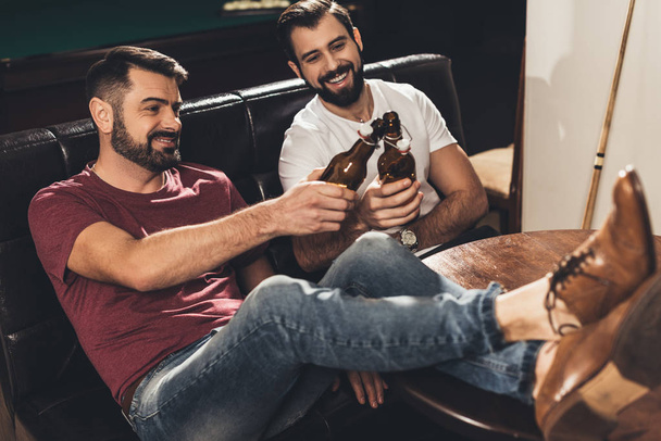 hombres caucásicos jóvenes sentados en el sofá y bebiendo cerveza junto a la mesa de billar en el bar
 - Foto, imagen