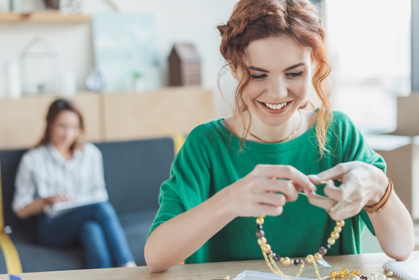 glückliche junge Frau macht Halskette aus Perlen in der Werkstatt, während ihr Kollege auf dem Sofa sitzt verschwommen auf dem Hintergrund - Foto, Bild