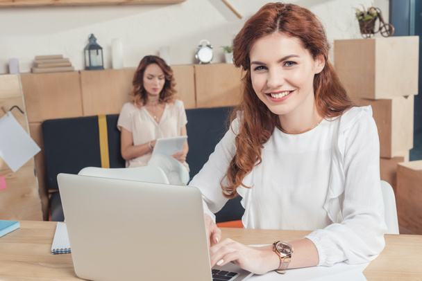joven mujer de negocios que trabaja con el ordenador portátil en la oficina mientras que su colega sentado en el sofá y el uso de la tableta
 - Foto, imagen