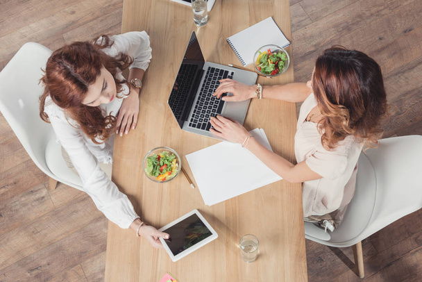 вид на бизнес-женщин, обедающих вместе и использующих устройства в офисе
 - Фото, изображение