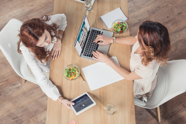 Blick von oben auf junge Geschäftsfrauen, die gemeinsam zu Mittag essen und Geräte im Büro benutzen - Foto, Bild