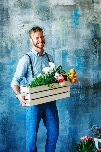 Male florist holding wooden box with flowers on blue background - Fotografie, Obrázek