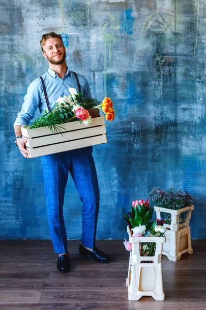 Male florist holding wooden box with flowers on blue background - Foto, Bild