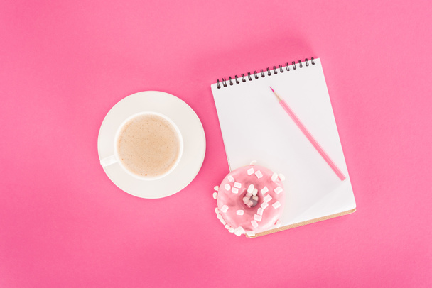 bovenaanzicht van geglazuurde donut met kopje koffie en lege notebook op roze oppervlak - Foto, afbeelding