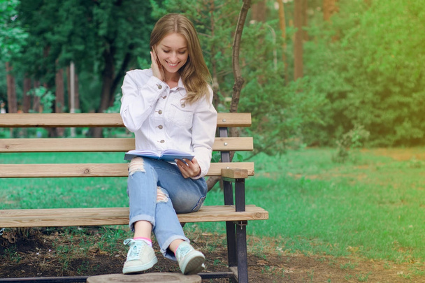 Beautiful young woman on a nature bench reading a book - 写真・画像