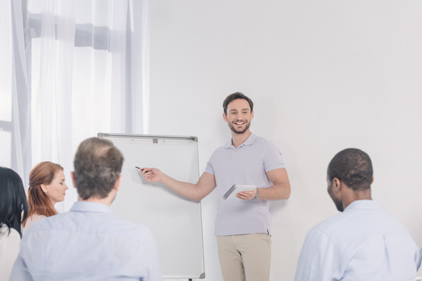 smiling man holding notebook and pointing at blank whiteboard while looking at multiethnic people during group therapy - Photo, Image