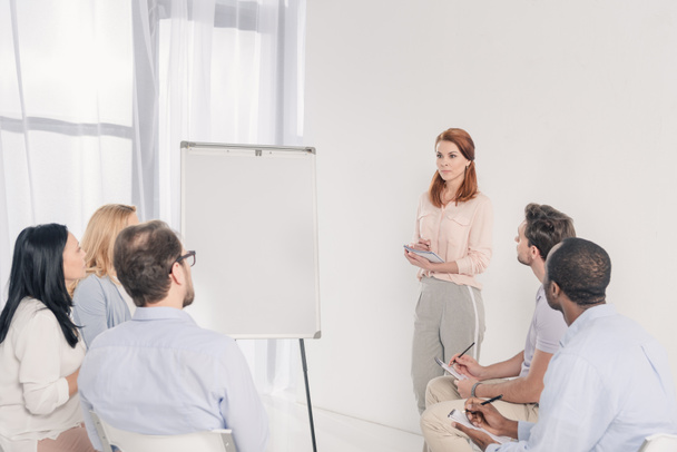 middle aged woman standing near blank whiteboard and looking at multiethnic people during group therapy - Photo, Image