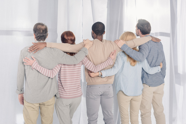 Rückansicht multiethnischer Menschen, die während der Gruppentherapie stehen und sich umarmen  - Foto, Bild