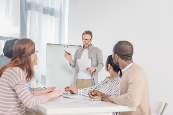 middle aged man in in eyeglasses holding paper and having conversation with his team while standing at whiteboard  - Zdjęcie, obraz