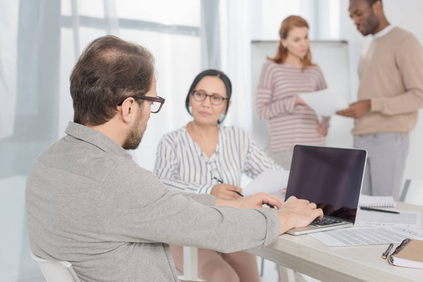 mature man using laptop with blank screen while multiethnic people sitting and standing behind - Photo, Image