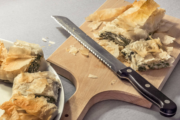 Torta de rolo de espinafre de queijo sérvio tradicional recém-assada cortada com faca serrilhada em placa de corte de madeira e servida em prato de porcelana
 - Foto, Imagem