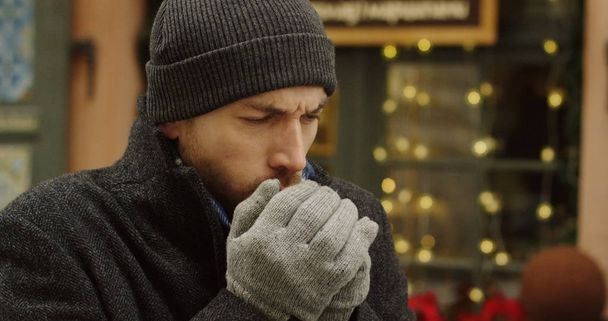 Portrait of the caucasian man in a hat and gloves coughing hard on the street in winter. Christmas decorated showcase behind him. Close up. Outdoor - Zdjęcie, obraz