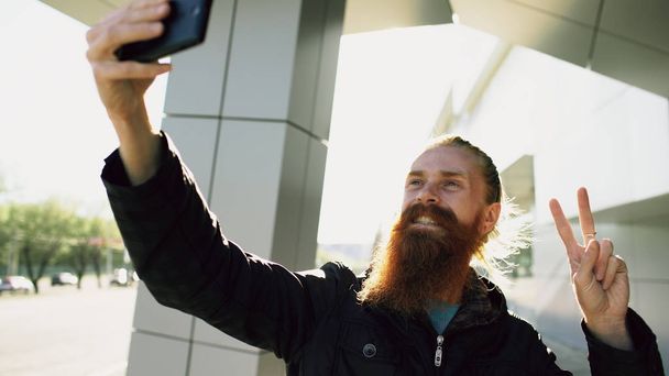 Young bearded hipster man taking selfie picture using smartphone camera outdoors at city street - 写真・画像