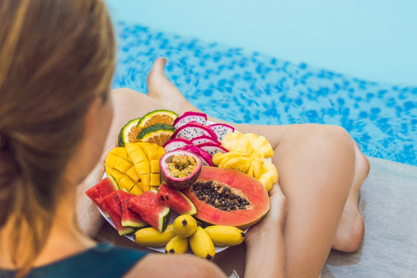 Young woman relaxing and eating fruit plate by the hotel pool. Exotic summer diet. Photo of legs with healthy food by the poolside, top view from above. Tropical beach lifestyle. - Фото, изображение