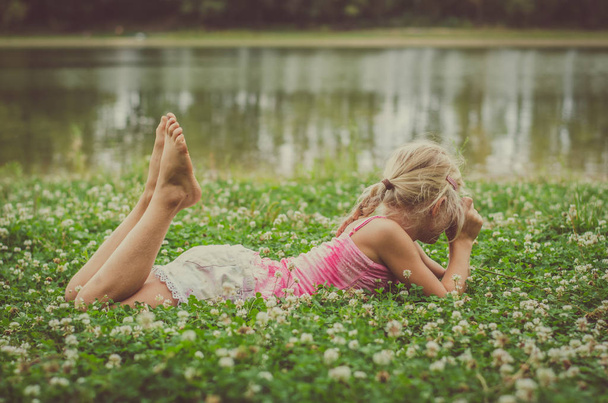 menina encantadora com cabelo loiro deitado na grama junto ao rio relaxante
  - Foto, Imagem