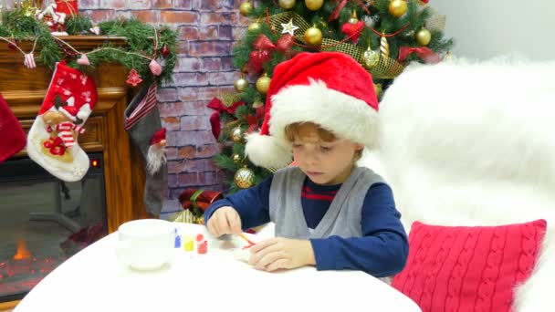 Um menino de chapéu de Pai Natal está pintando um brinquedo de Natal
. - Filmagem, Vídeo