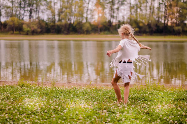 маленька дівчинка з довгим світлим волоссям у білій сукні танцює в зеленій траві біля річки
 - Фото, зображення